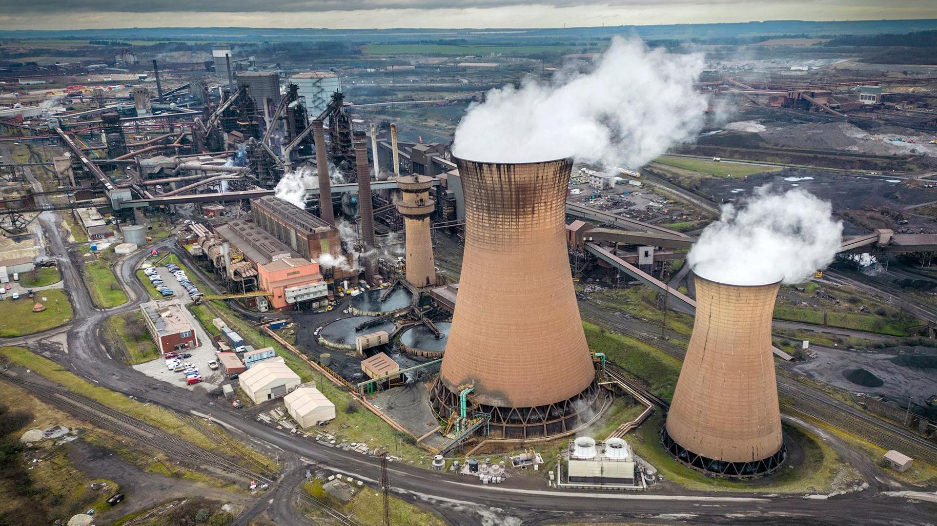 Steam comes out of huge towers in a wide shot of Scunthorpe steel works against a cloudy sky in 2023.