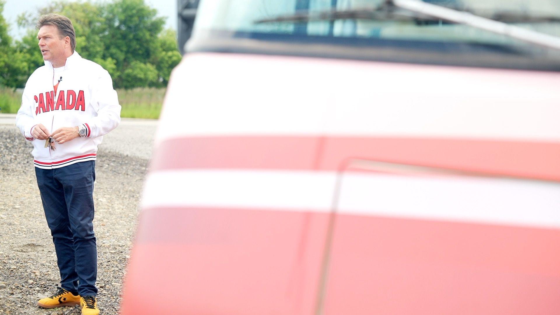 Thomas Lukaszuk stands next to his red and white Unity Bus in Calgary. He is wearing a white zip up fleece jacket with big red letters on the front that read CANADA, dark blue jeans and yellow and black sneakers. He has short dirty blonde hair.