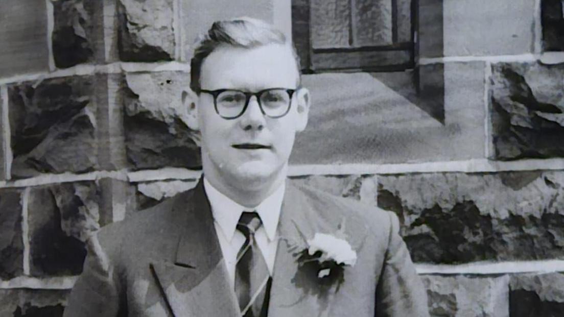 A black-and-white head-and-shoulders photograph of Anthony Littler, a middle-aged man with swept-back hair and thick dark-framed glasses, wearing a dark suit jacket, shirt and tie, with a slight smile.