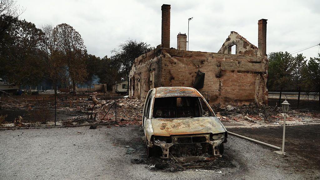 Burned vehicles, damaged buildings, destroyed roads, and charred trees are seen across areas affected by the blaze.