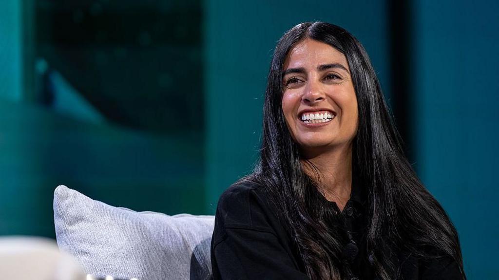 A woman with long dark hair sits on a sofa on-stage at a business event. She looks relaxed as she smiles at the host off-camera.