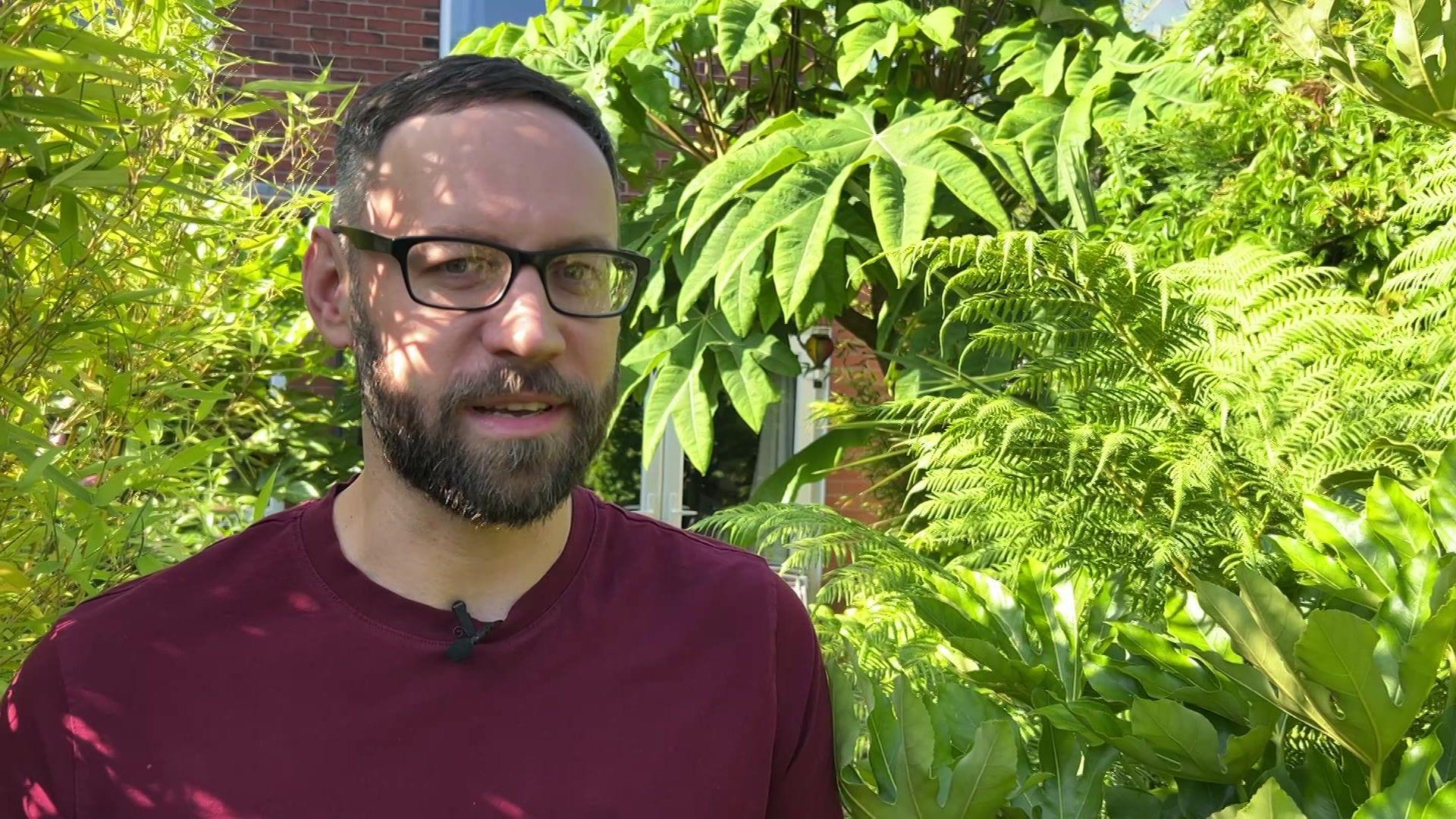 A picture of Dave Iveson who has dark brown hair, glasses and a beard standing in his garden. He is surrounded by lots of tropical plants and trees.