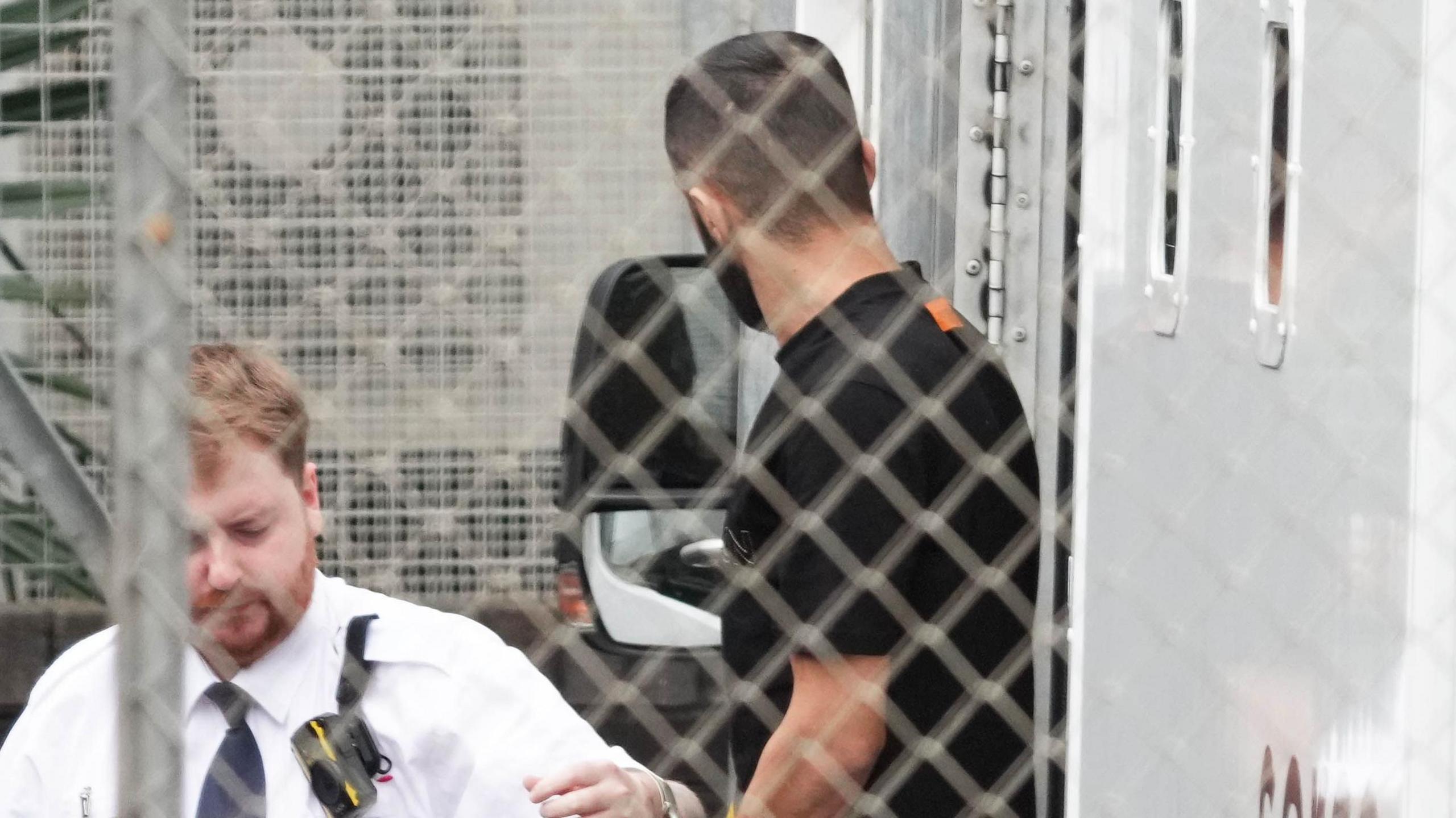 A man in a white uniform leads a man in dark clothing, who he is handcuffed to, out the side of a white prison van. They are pictured behind a grey wire fence.