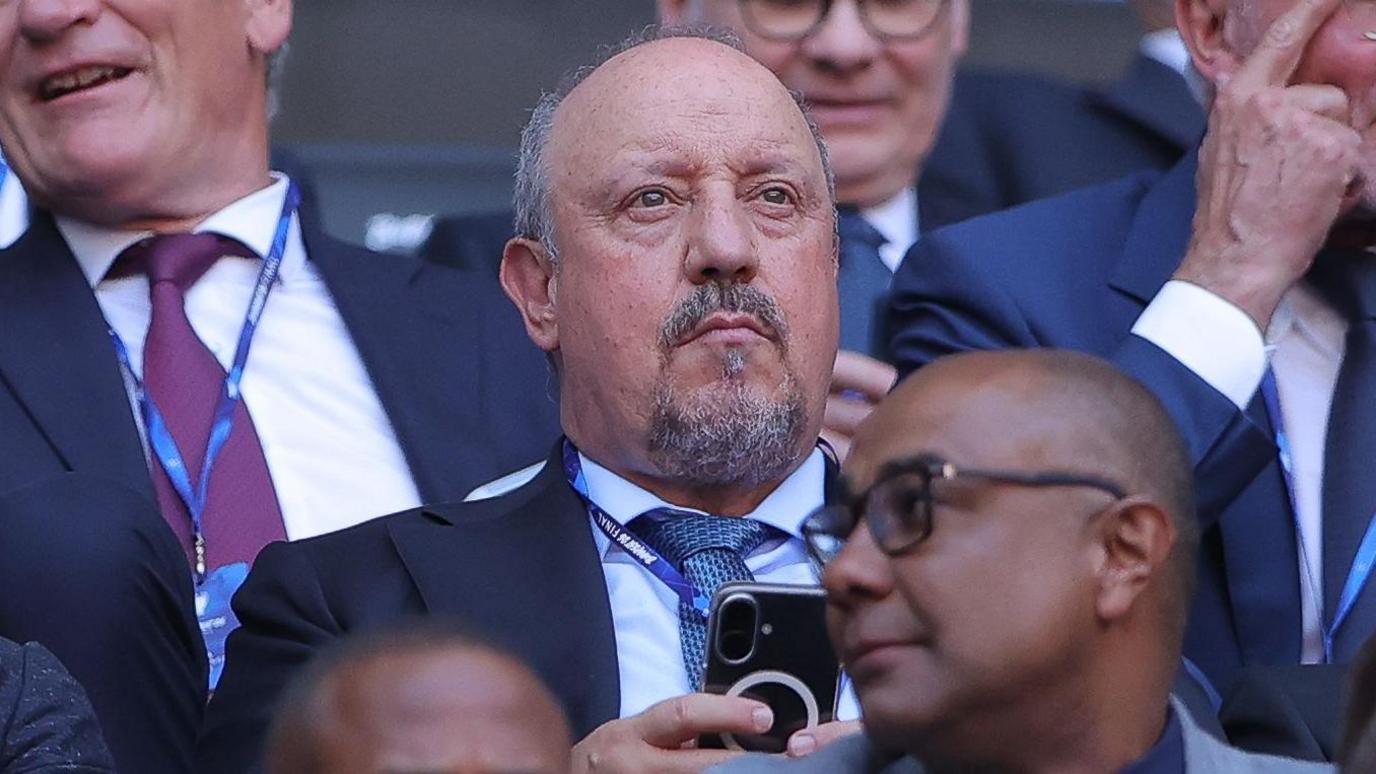 Rafael Benitez, wearing a suit and tie, holds his phone with two hands while watching a match