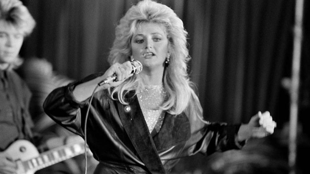 Black and white image of Bonnie Tyler, with big blonde hair, singing into a microphone. She is wearing a leather trench coat, with a musician playing a guitar in the background.