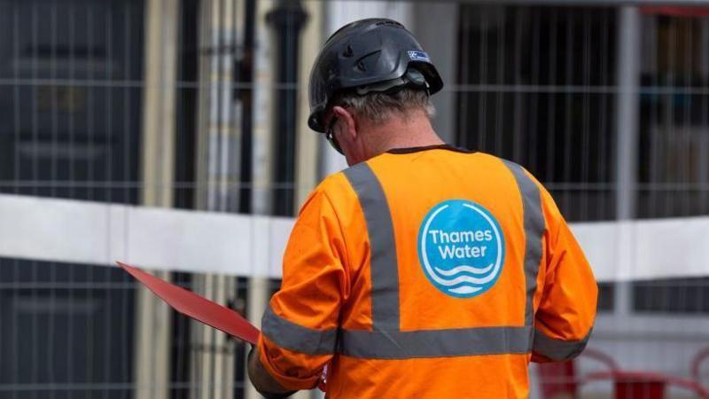 Male Thames Water worker seen from behind, wearing an orange high-vis jacket, with Thames Water's blue logo on the back, a black hard hat and looking at an open red folder in his hands