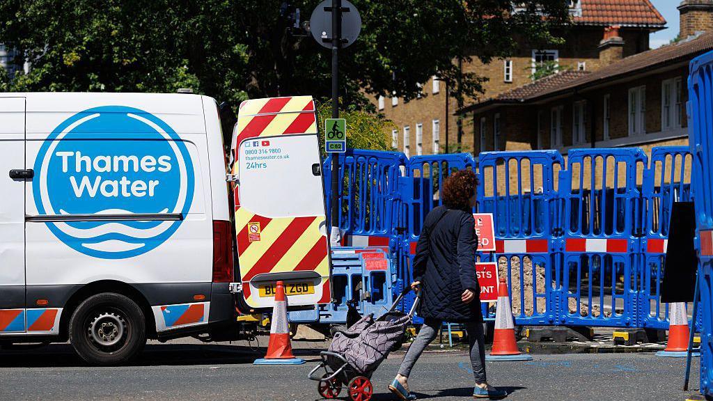 The side of a Thames Water van with an open back door parked on a residential street next to a blue fence blocking off some work it is doing. A woman in the foreground with a blue coat, grey leggings, and a backpack on wheels walks past the scene.