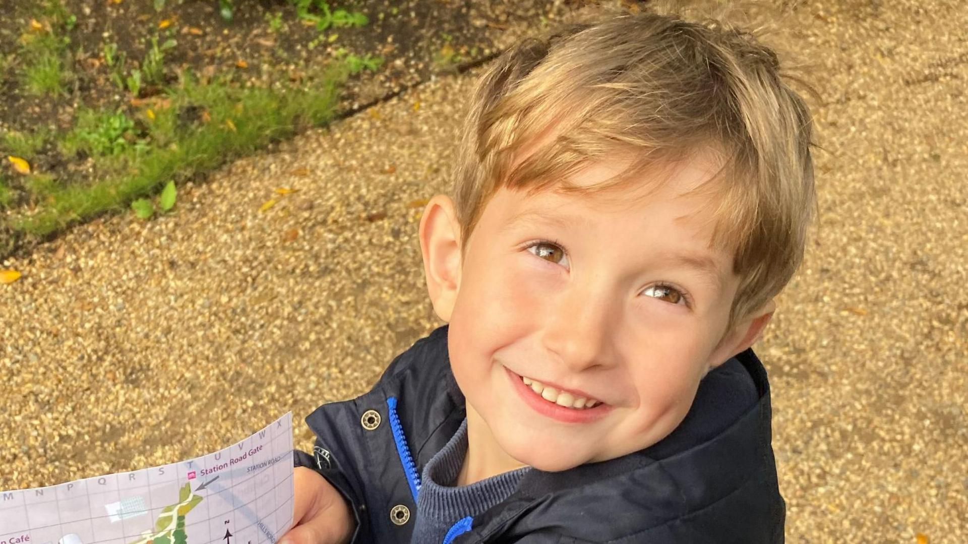 Benedict, a young boy with blond hair, on a path holding a map. He is wearing a black coat and is smiling to the camera.