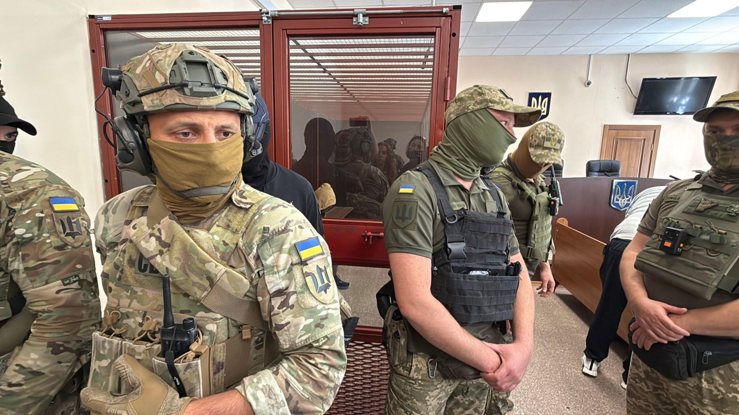 Men in military fatigues stand in a courtroom with a person sitting in a glass room