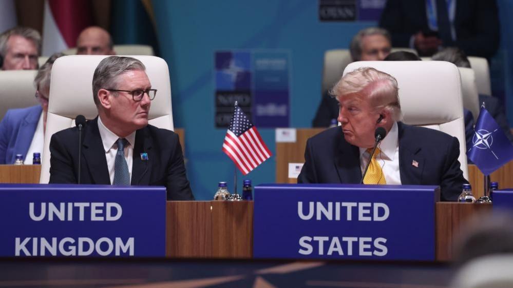 Sir Keir Starmer and Donald Trump are sat down in white chairs and looking at each other. There is a miniature United States flag between them on a wooden table and a miniature blue flag with the Nato emblem next to Trump's left arm on the table. Both men are wearing dark coloured suits, white shirts and ties.