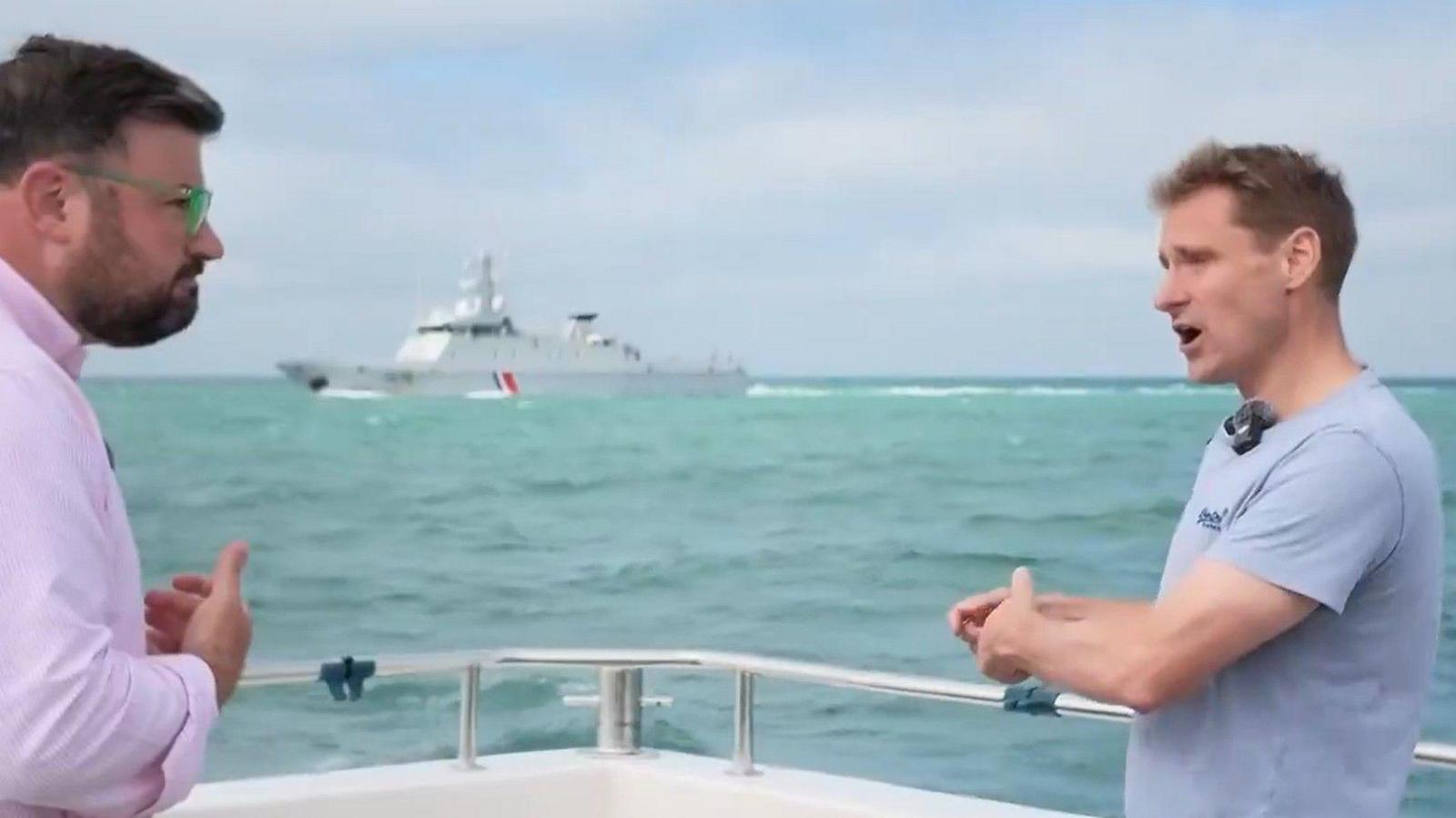 MP Chris Philip, in a grey t-shirt, stands on a boat in the sea, speaking to a man with glasses in a pink shirt. A large, grey ship is visible in the background.