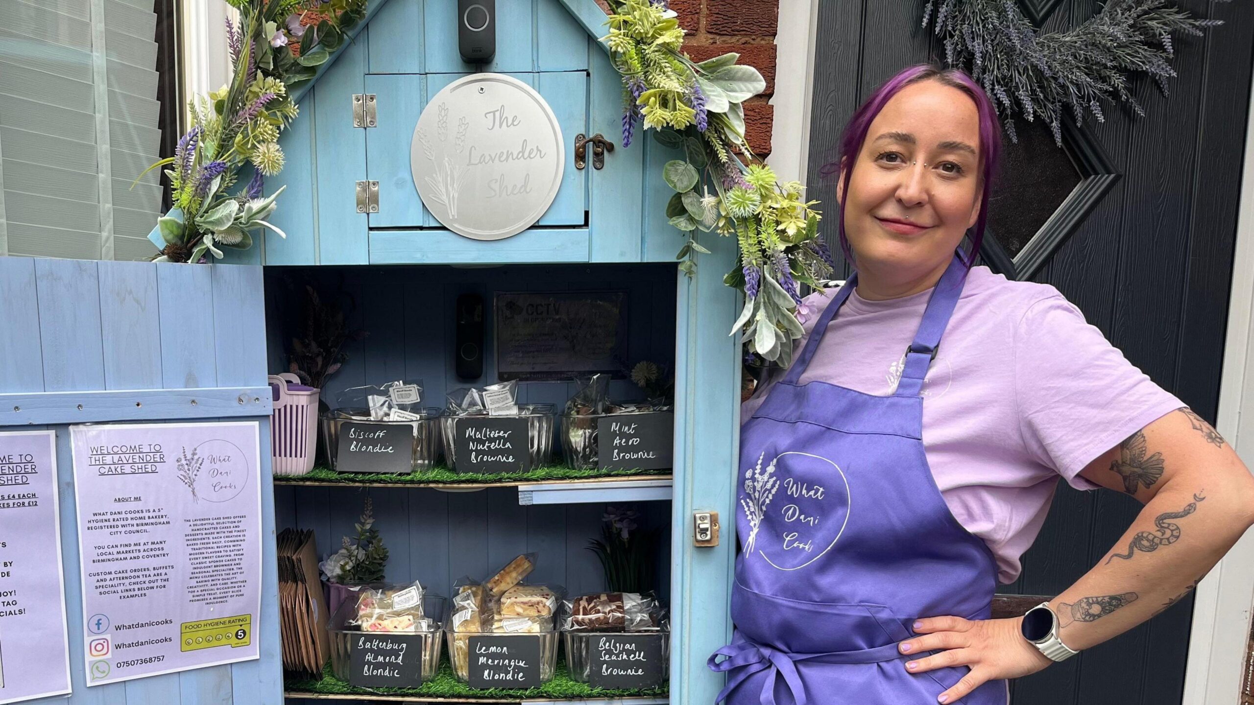 Dani wearing a purple apron and purple t-shirt standing next to the cake shed. The door is open and there are three shelves of cakes. The shed is blue and has a floral garland on the top