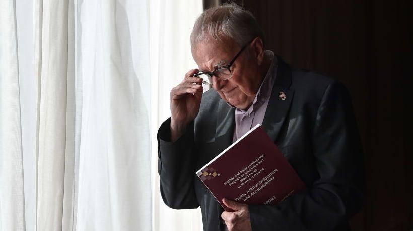 Professor Phil Scraton is seen clutching a report, he has white hair and is holding his glasses.