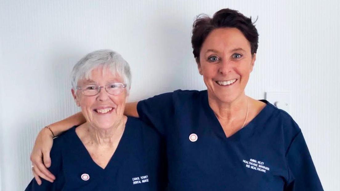 A portrait shot of Carol and her daughter Emma. Both are dental nurses and wearing blue uniforms. Emma is hugging her mother and has her arm around her.