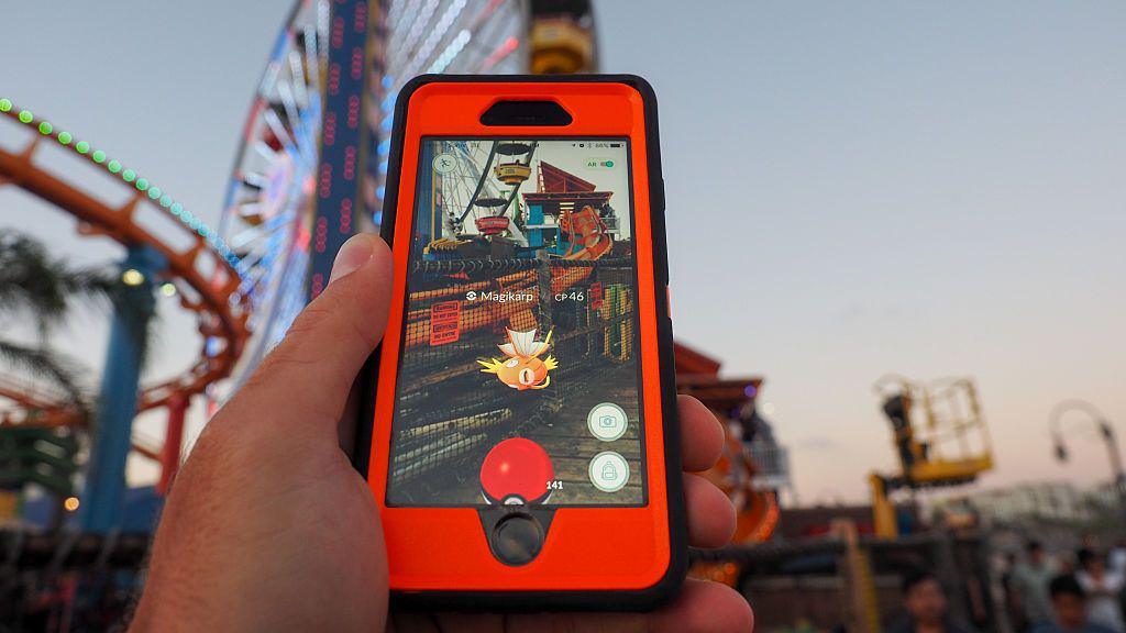 A red and black phone showing the camera view of a location with an augmented Magikarp from Pokémon Go. In the blurred background there is a ferris wheel and a rollercoaster track.
