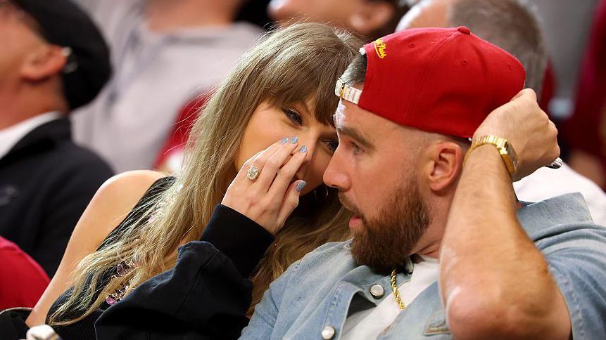 Close up of Taylor Swift whispering behind her hand to Travis Kelce who is wearing a red cap pointed backwards