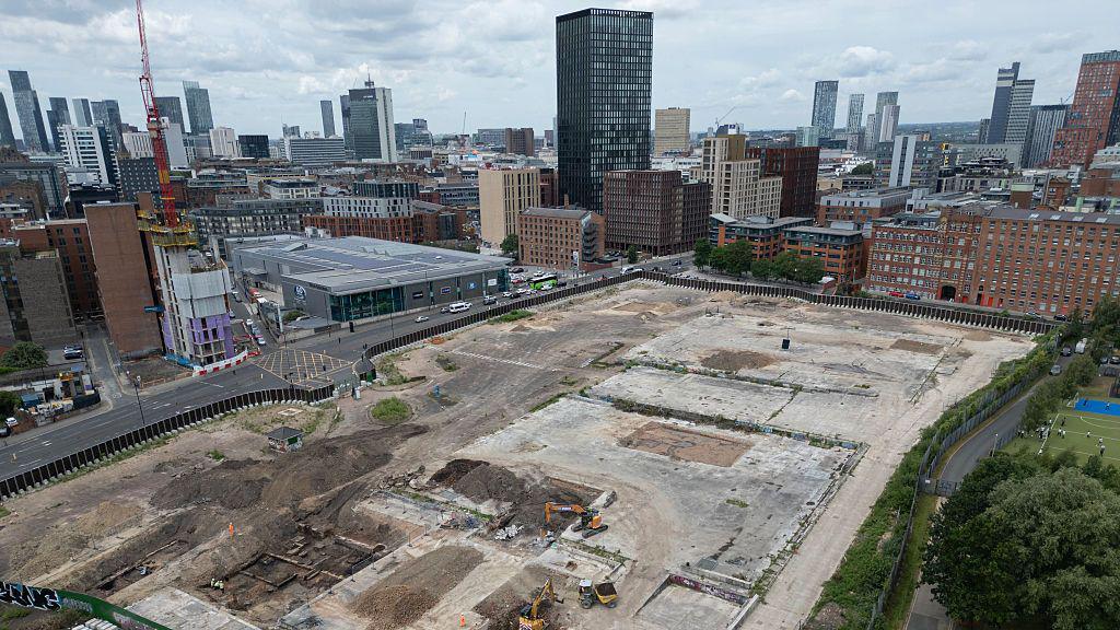 An aerial shot of two diggers in the middle of a large piece of empty land. There are buildings on the edges of the land, as well as an area of green space. In the background, there are clusters of high-rise towers.