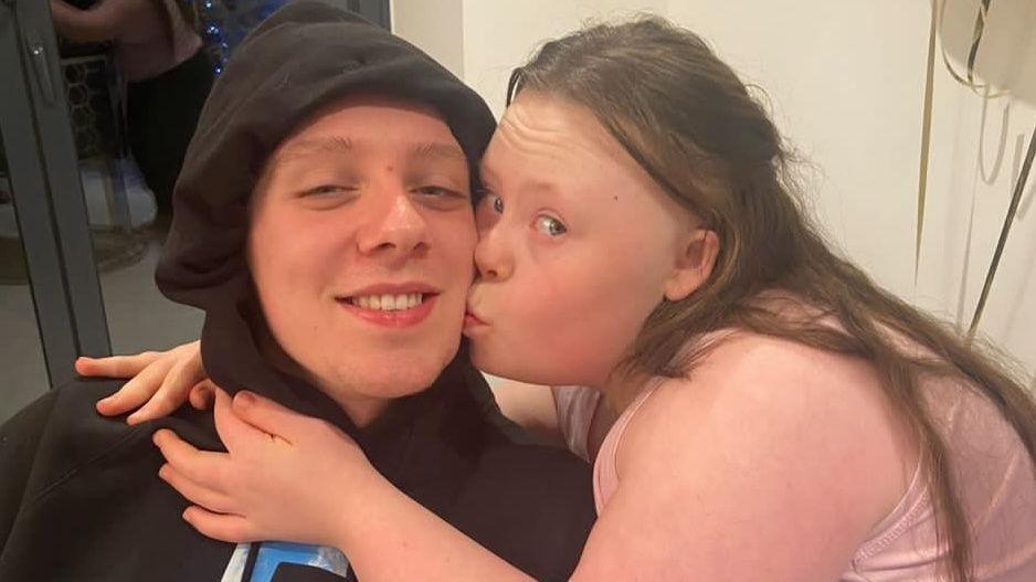 Aitch posts with his sister Gracie, who is kissing him on the cheek. Aitch is wearing black hoodie with the hood up. Gracie is wearing a pink top.