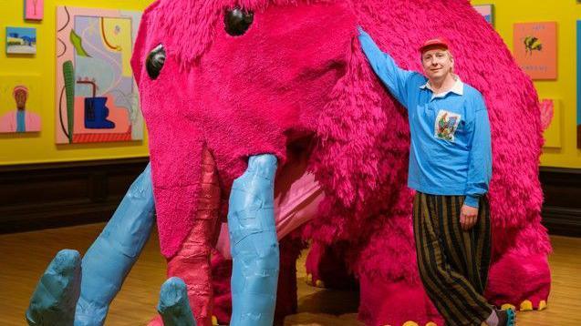 A man in striped black and beige trousers stands his arm resting on a life-size sculpture of a woolly mammoth, with pink fur, blue horns and yellow toenails. He is in an art gallery with yellow walls, with paintings hung.