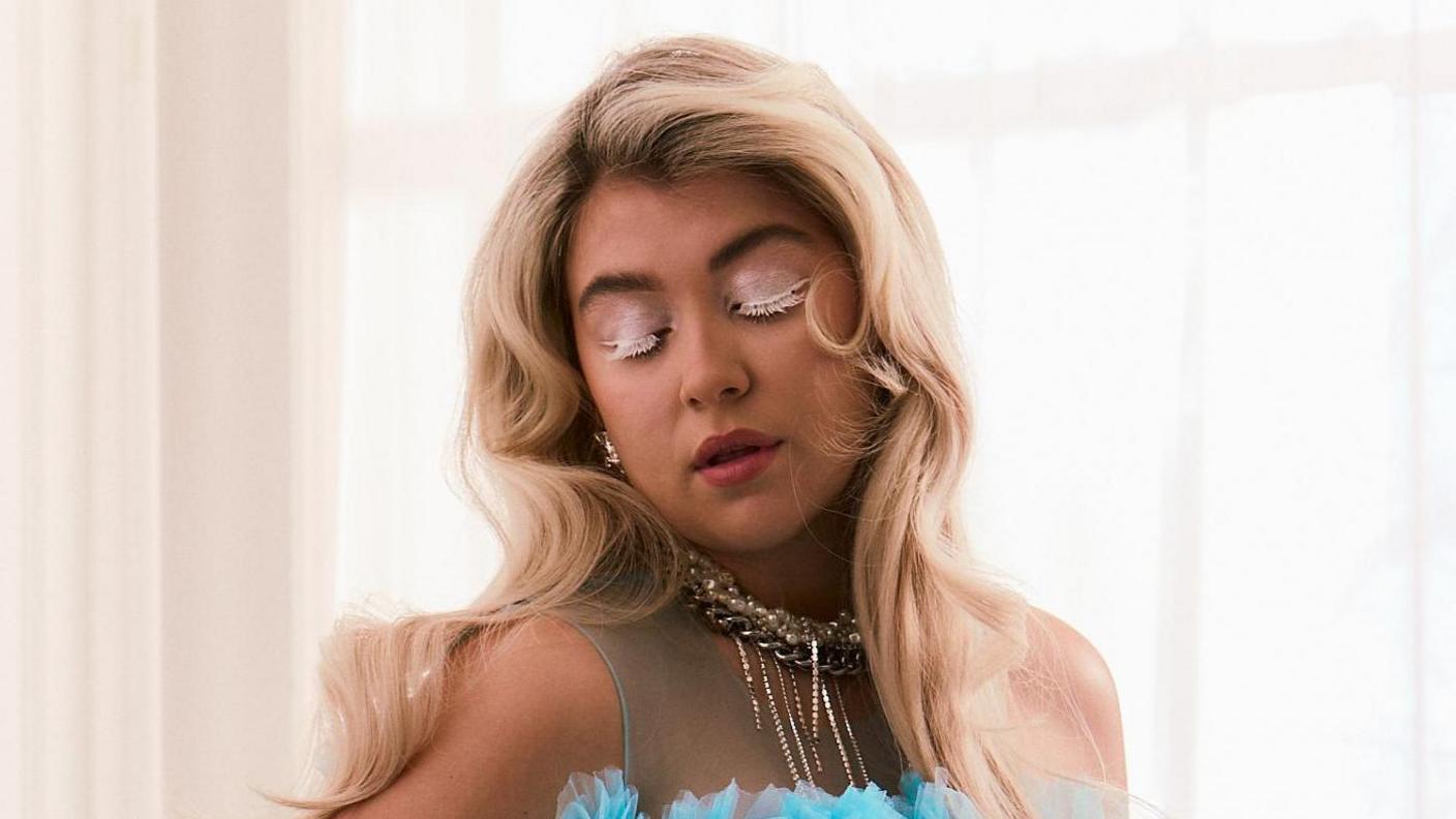 Caity Baser in a promotional shot for her new single. She has long blonde hair worn loose and wears a bright blue tulle dress with a statement gold necklace. She wears red lipstick and white mascara and looks down. She's pictured in a bright, airy room with net curtains covering a sunny window behind her.