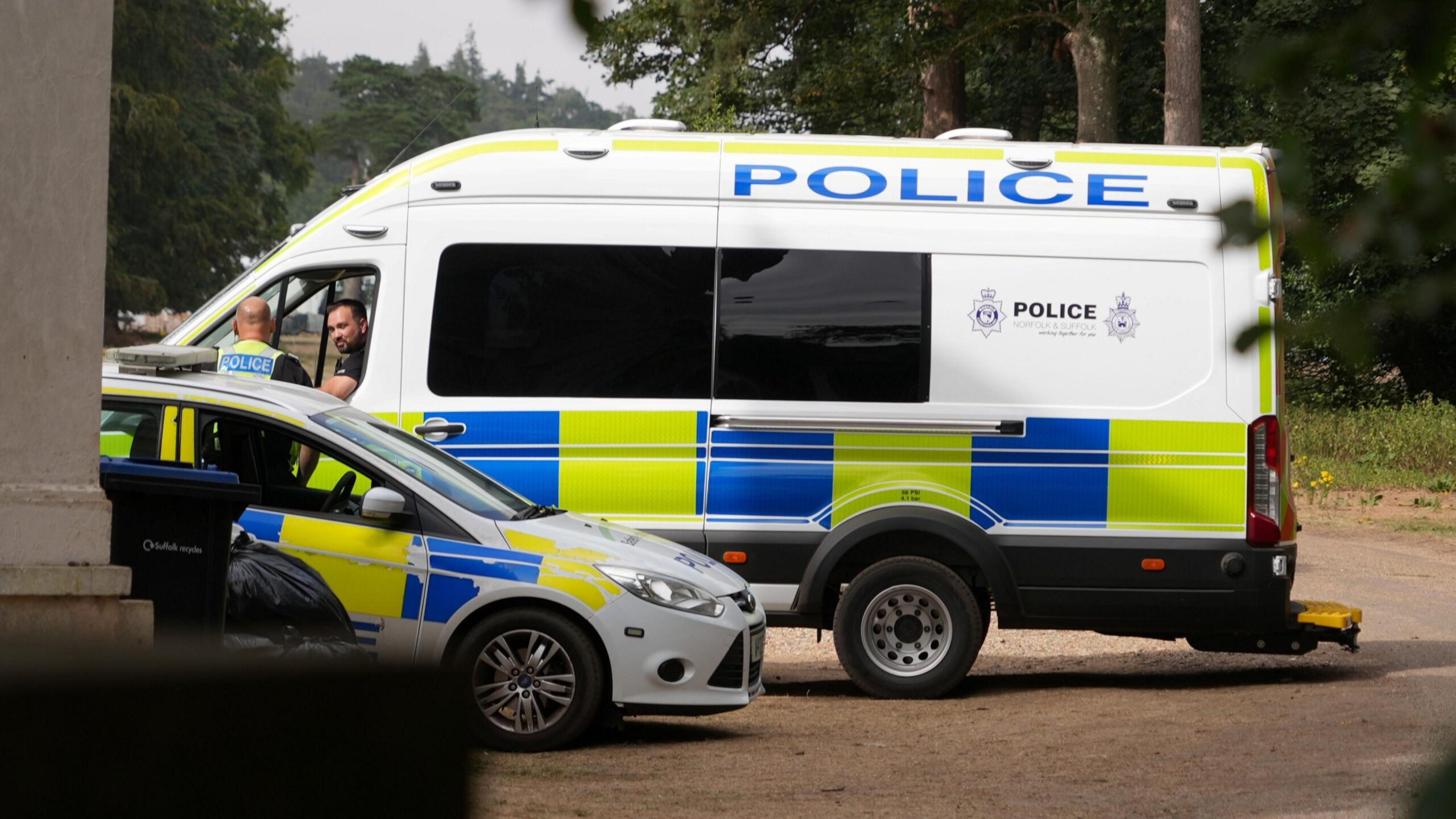 A large police van and a police car parked next to each other. Two officers - including one in the passenger seat and one standing outside - talking