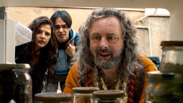 A man with long, curly grey hair and facial hair, staring into a cupboard full of jars. A young man and young woman, both brunette, are stood behind him, also leaning down to look at what he is looking at. They have puzzled looks on their faces.