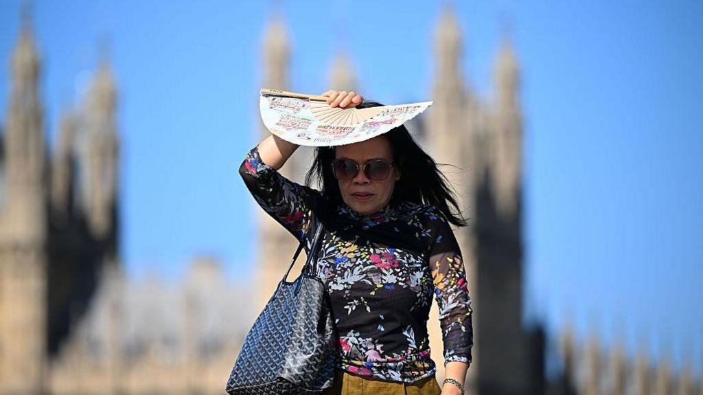 A woman in bright sunlight shades her face with a patterned paper fan