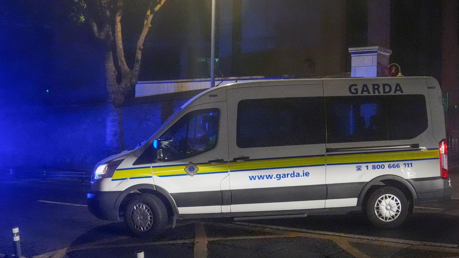 A white Garda van with a yellow stripe along its side pulls out onto a main road on a dark night.