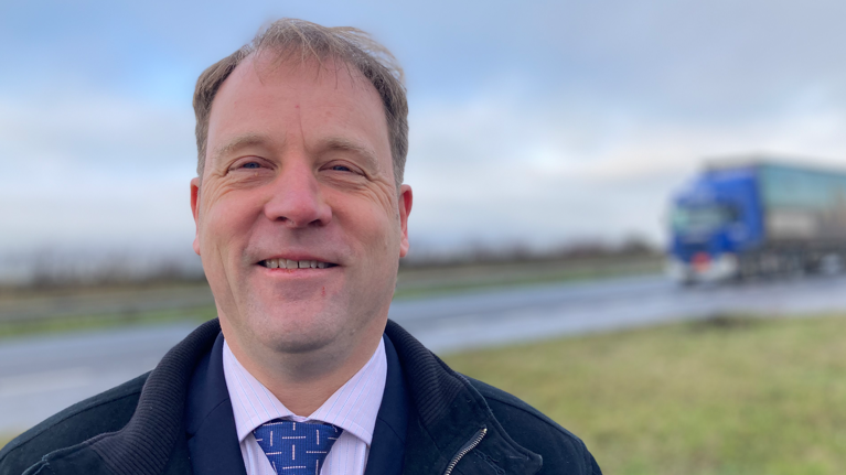 A man wearing a blue jacket and a blue tie stands in front of a road. He has receding light brown hair and is smiling at the camera. There is a HGV entering the picture in the background.