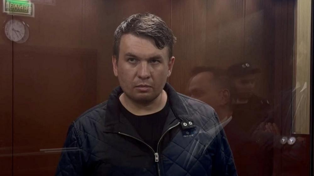 A darkened photo of a man in court wearing a dark jacket, standing behind a glass screen