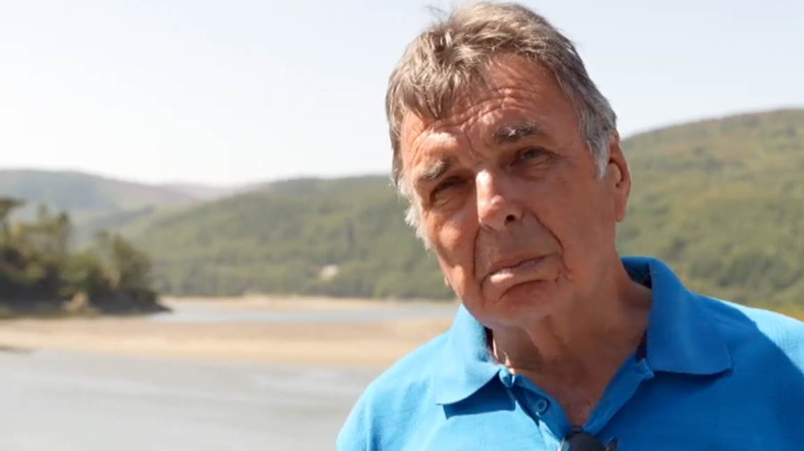 Man with blue polo shirt and greying temples looks towards camera, with sandbanks of an estuary behind him, and rolling green hillsides