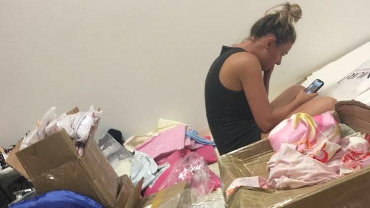 Olivia hunched over her phone with her hair in a bun surrounded by boxes of clothes.