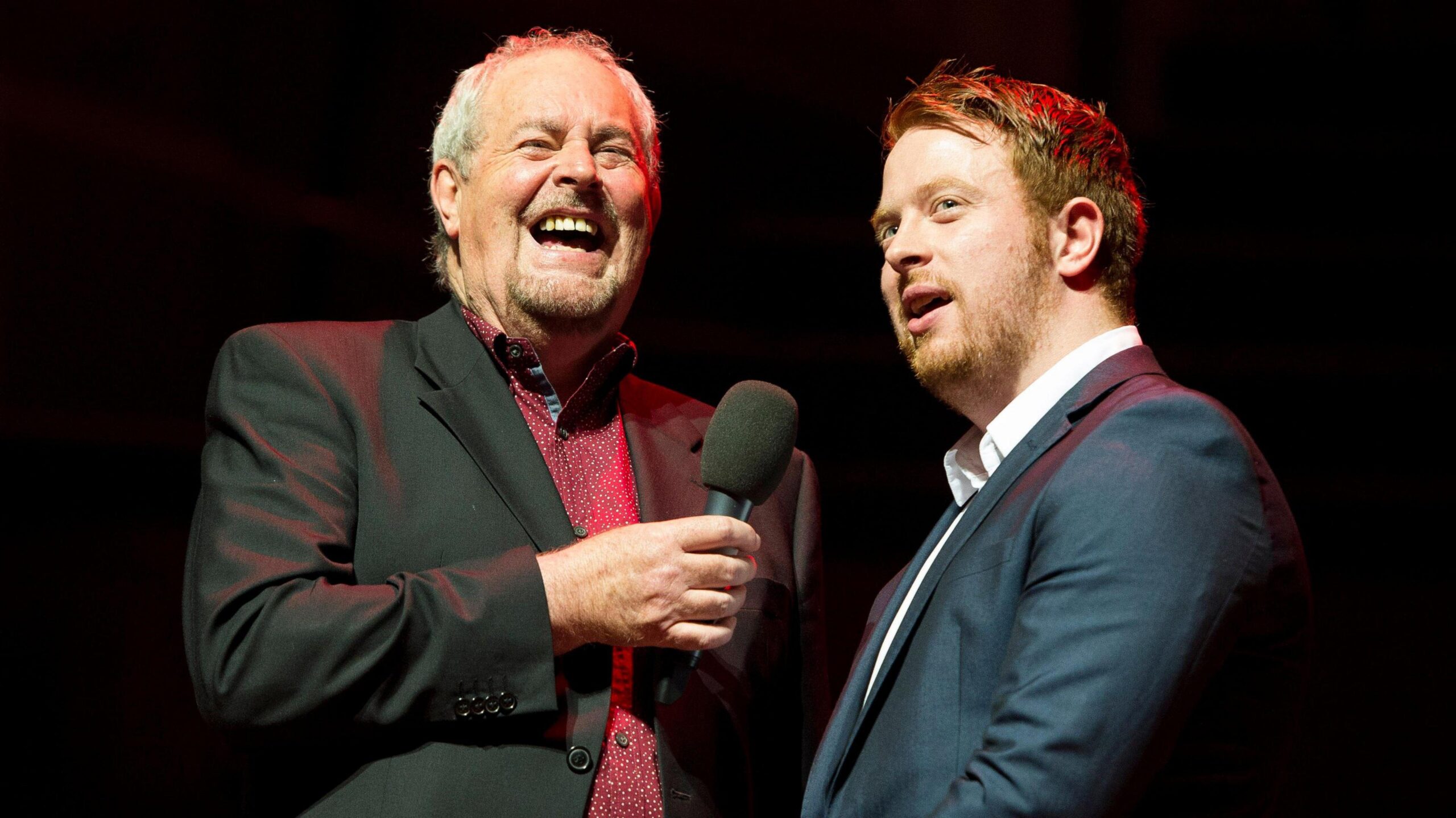 Two men standing side by side wearing shirts and blazers, one has short dark hair and one has short grey hair.