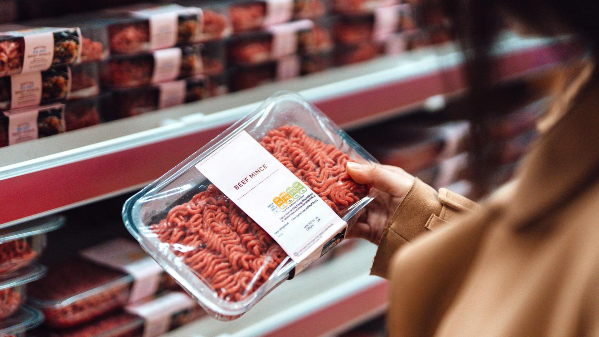 A woman holds a packet of beef mince in a supermarket with rows of mince on shelves in the background
