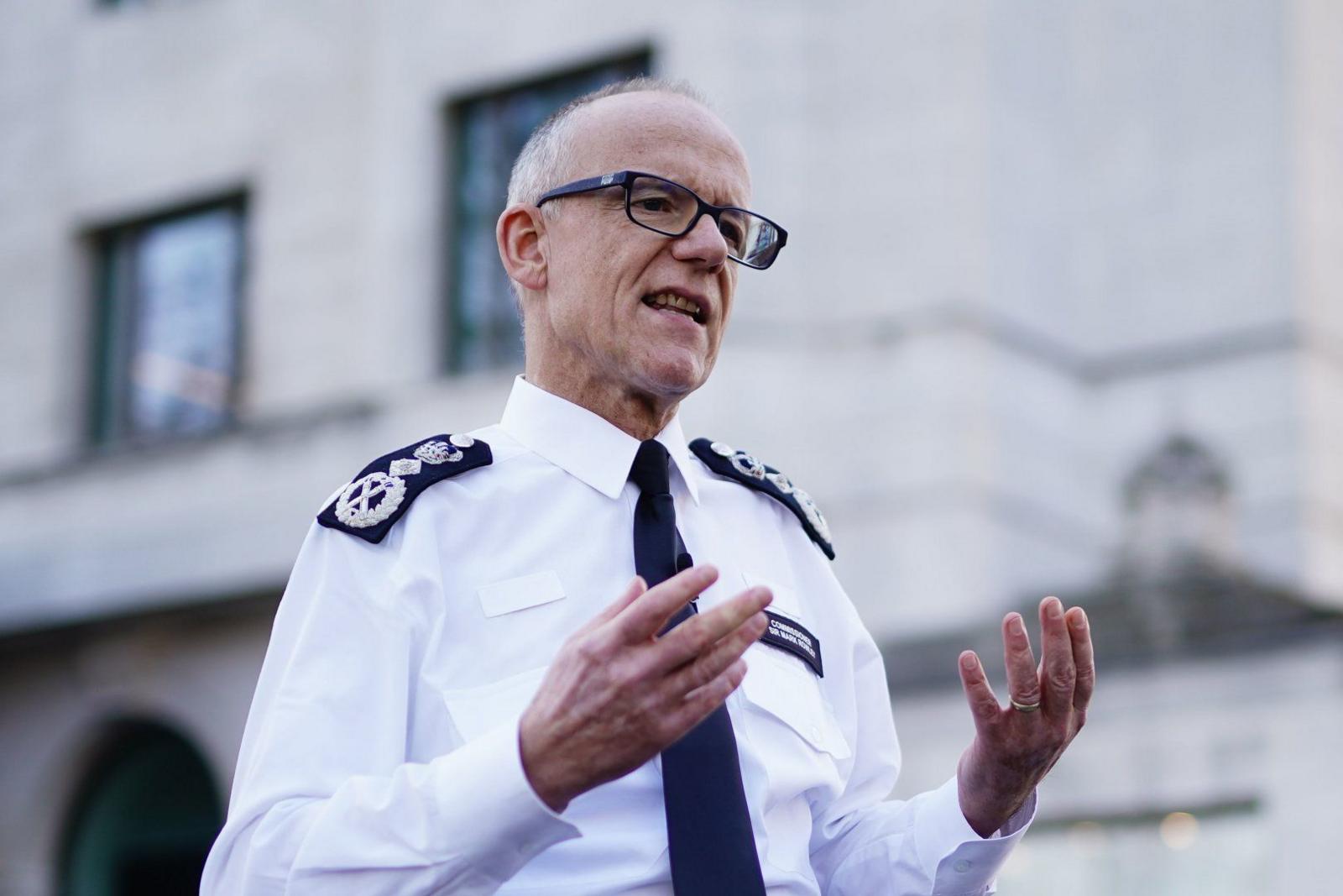 Sir Mark appears to be standing outside New Scotland Yard, addressing the media, he is wearing his police uniform and glasses and gesticulating with his hands.