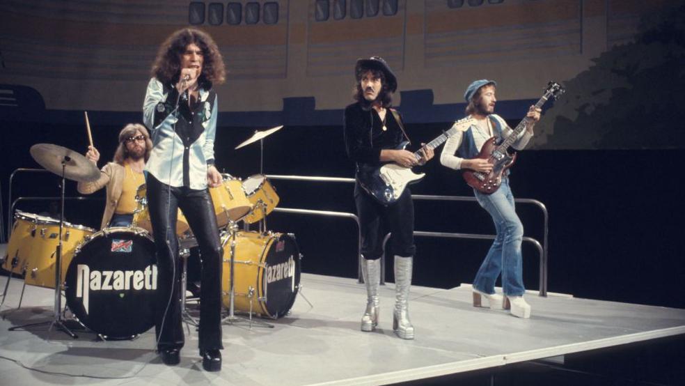 Nazareth are playing on stage in 70s attire, including leather trousers, platform boots and hats.