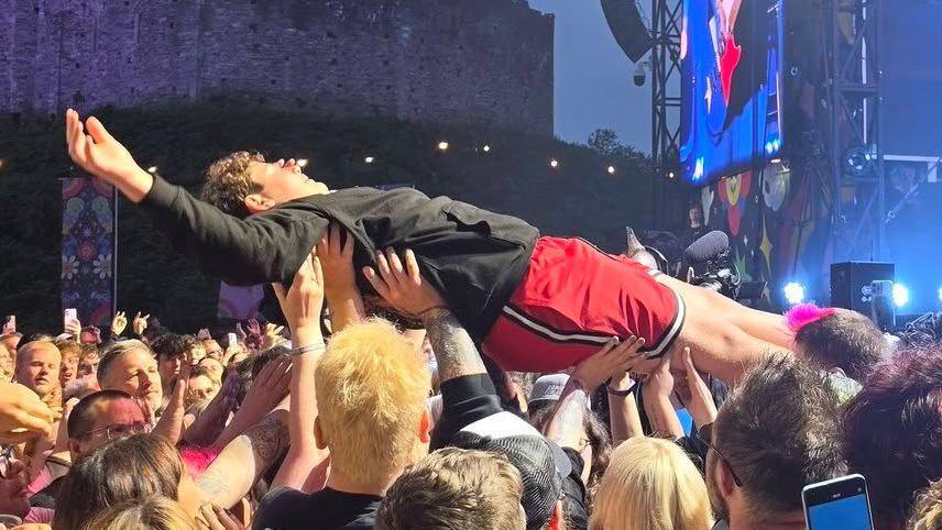Jaret Reddick son, Everett Reddick doing a crowd surf during the Bowling for Soup show at Cardiff Castle. Everett is pictured being heled up by fans, with his arms in the air. He is wearing a black hoodie and red shorts. Three team members (two men and one woman) can be seen standing by as he is lifted.