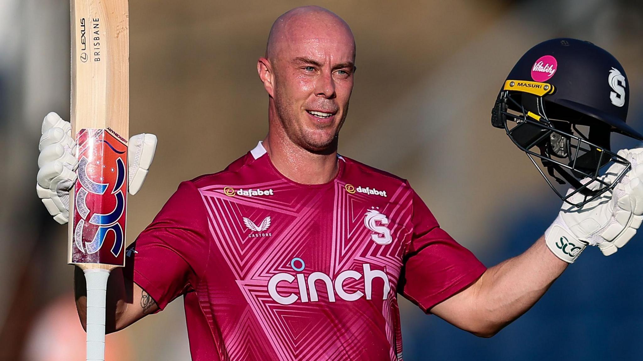 Chris Lynn, wearing Northamptonshire's red shirt, raises his bat and helmet to celebrate scoring a century against Glamorgan