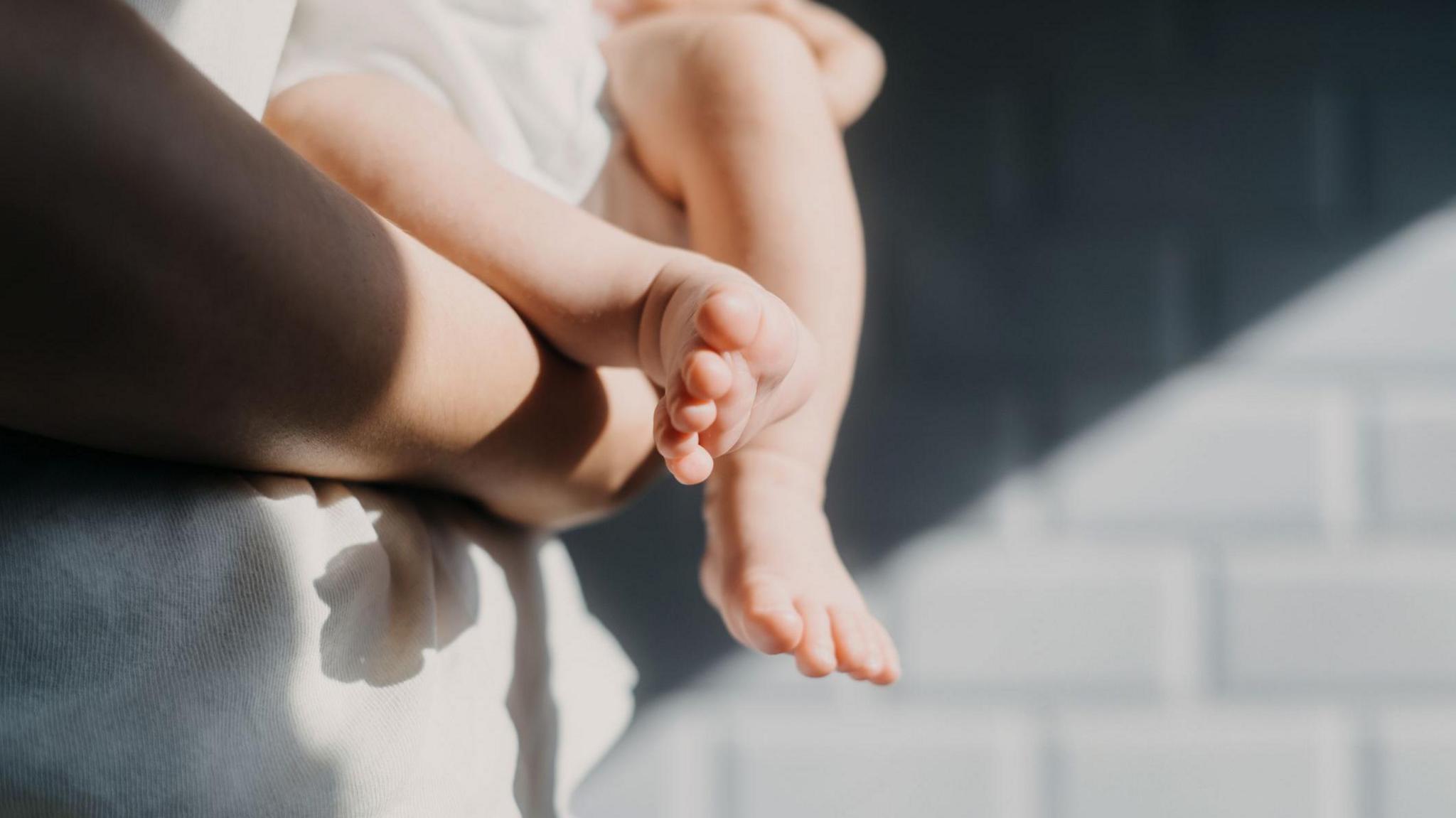 Close-up of a baby's feet. The baby is held by an adult wearing a white shirt, standing in front of a grey brick wall