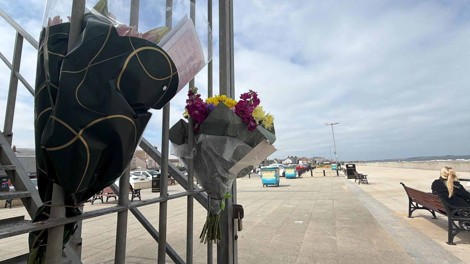 Side shot of a gate with two bouquets of flowers attached to it on a beach. The flowers are yellow and purple. The sky is blue.