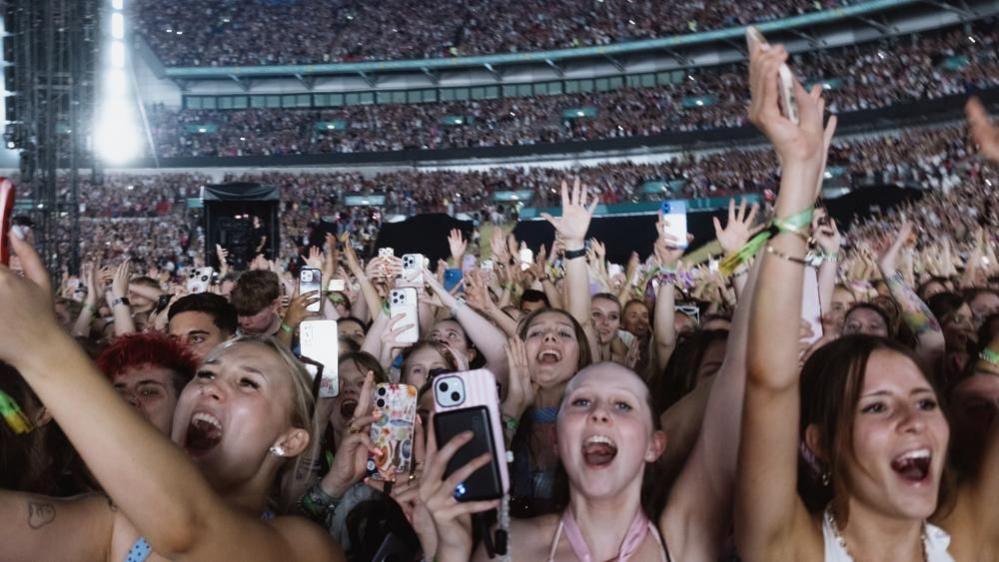 Several rows of predominantly female fans can be cheering, some holding their hands up, others holding their phones up in a shot of fans at Wembley Stadium for Harry Styles' performance last night. In the background, thousands of other people can be seen sat in the stands
