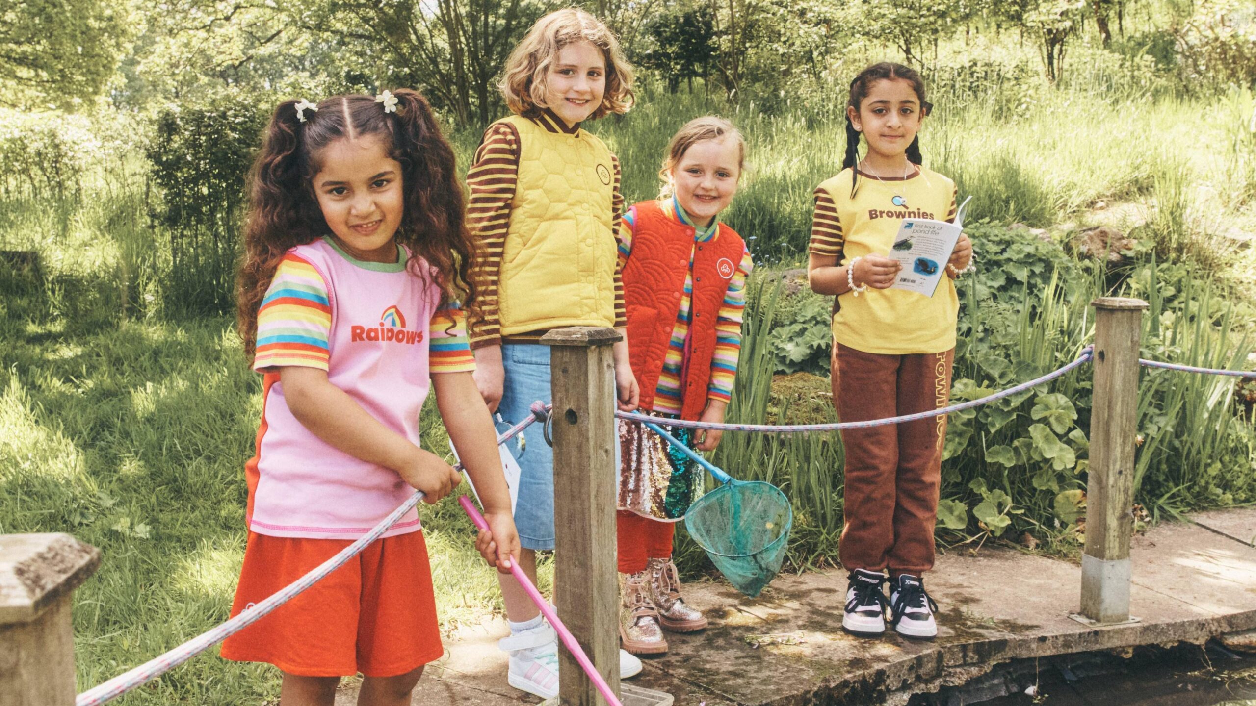 Four young girls, two Rainbows and two Brownies, standing by a pond in their new uniforms, with two girls holding nets.