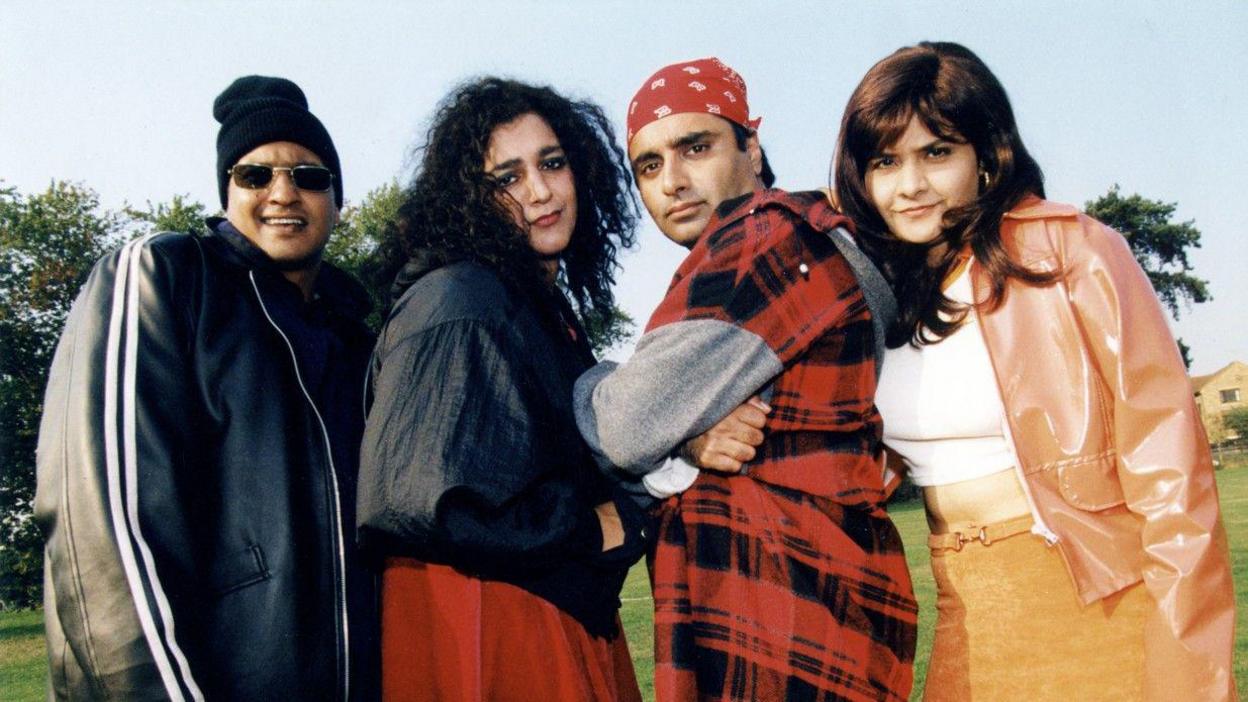 Four people are standing together on a grassy field with trees and a building behind them. They are posed in a straight line, each wearing different, brightly styled outfits.