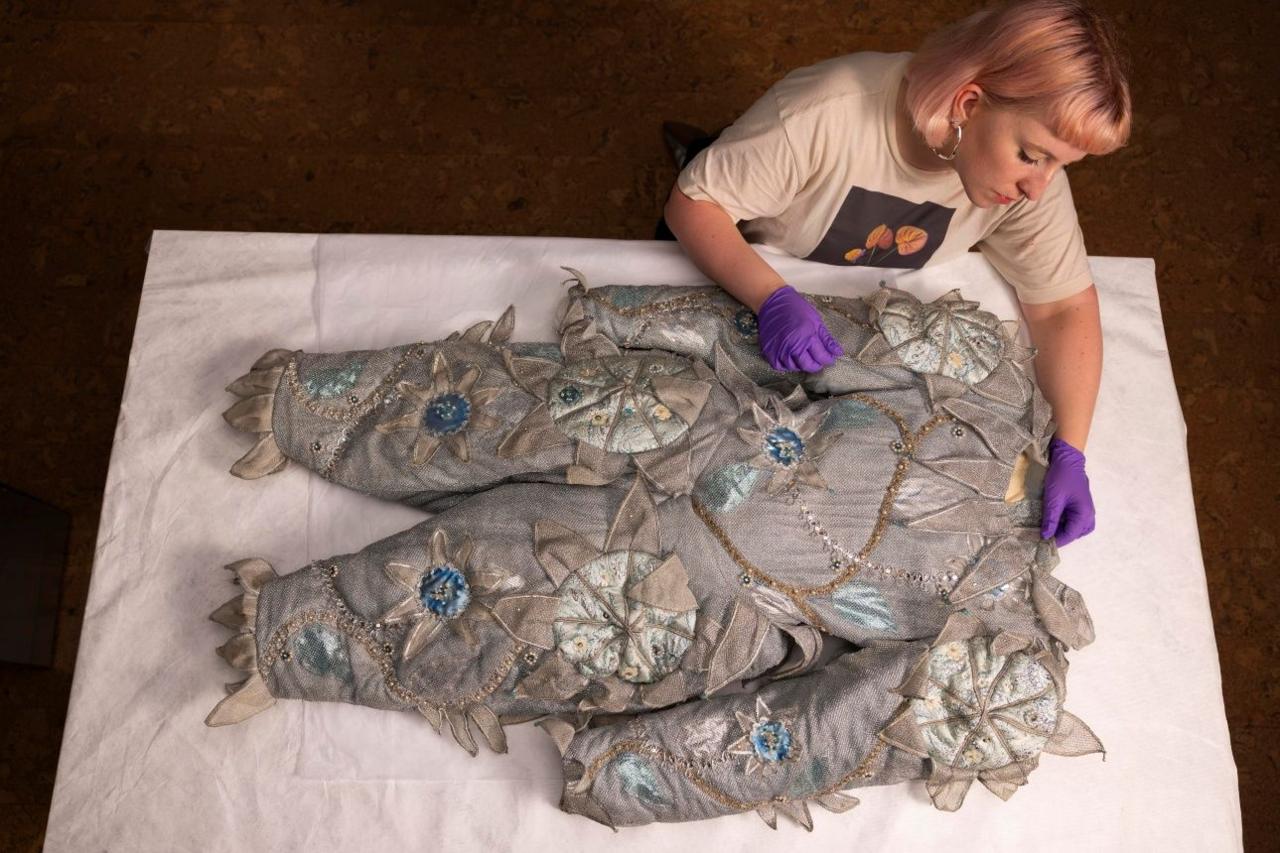 A grey clown's costume with flower patterns is displayed on a table with a museum curator examining the fabric
