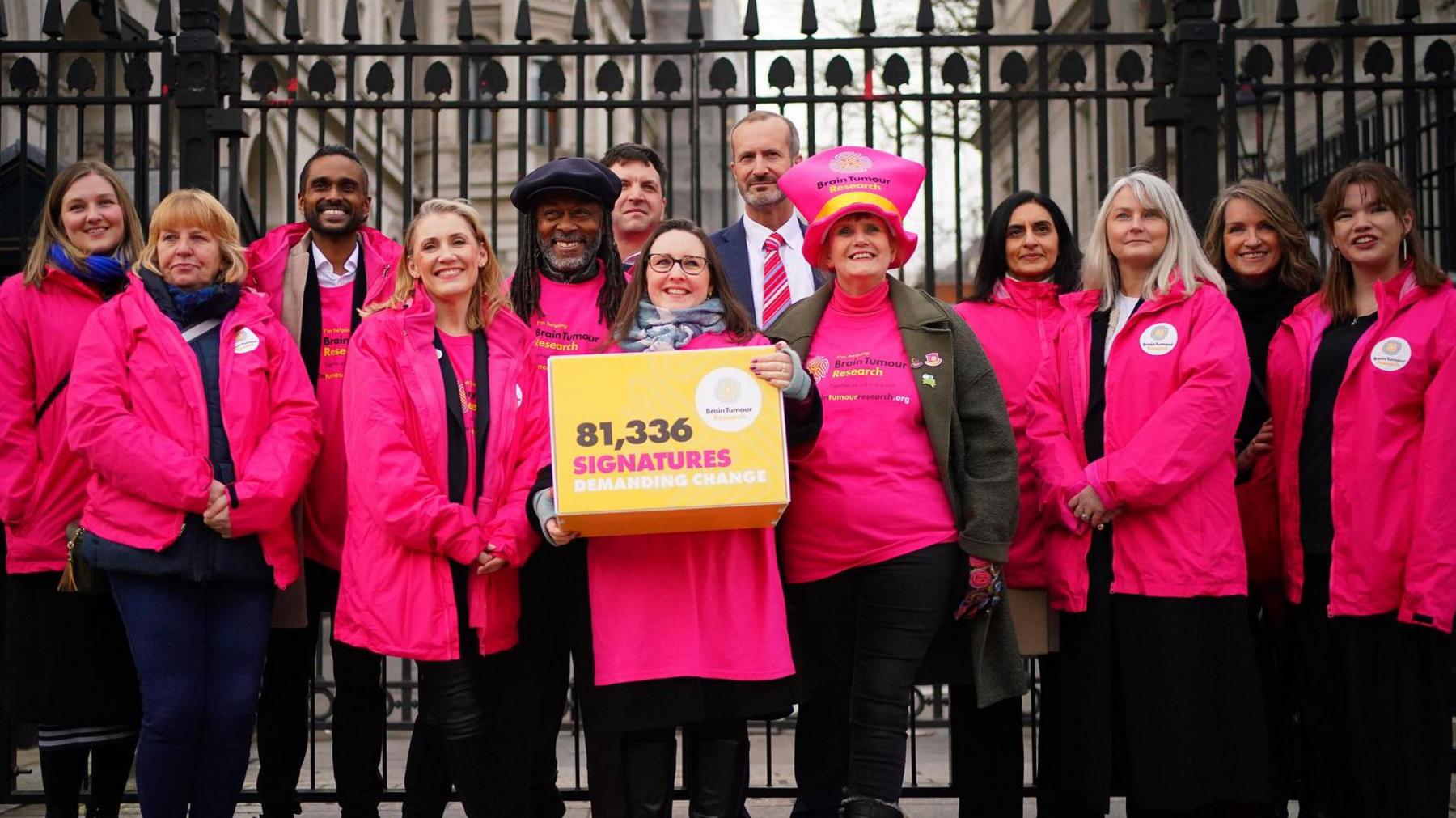 A group of 13 people stand by the gates for Downing Street wearing pink jackets and Theo Burrell is in the middle holding a yellow box that reads "81,336 signatures demanding change"