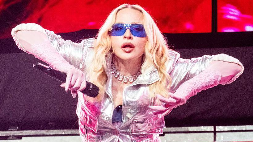 Madonna on stage in New York. She's holding her arms to the side, revealing pink lace gloves beneath a silver jacket. Her eyes are hidden behind jet blue sunglasses, but she appears to be staring directly at the camera.
