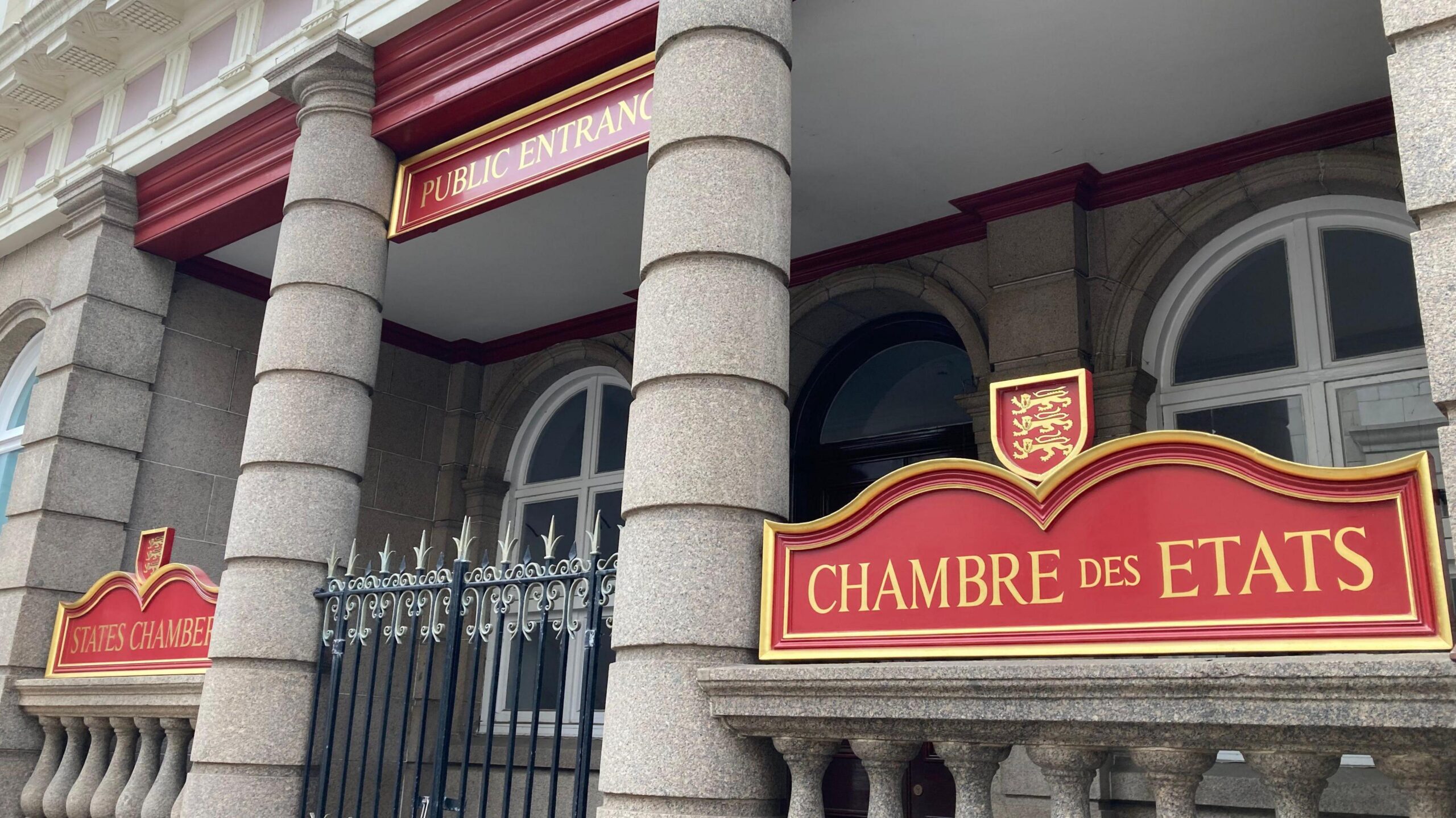 The front and public entrance of Jersey's States Assembly. The stone-fronted building has an outdoor foyer before its front doors, with two pillars and a metal gates. It also has red signs with gold lettering saying "States Chamber" and "Chambre Des Etats".