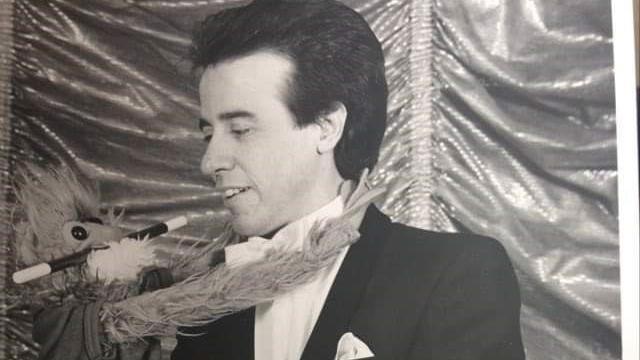 A black and white image of Reg Martin as a younger man performing magic. He is holding a plush doll with a magic wand in its mouth. Reg is looking at the doll.