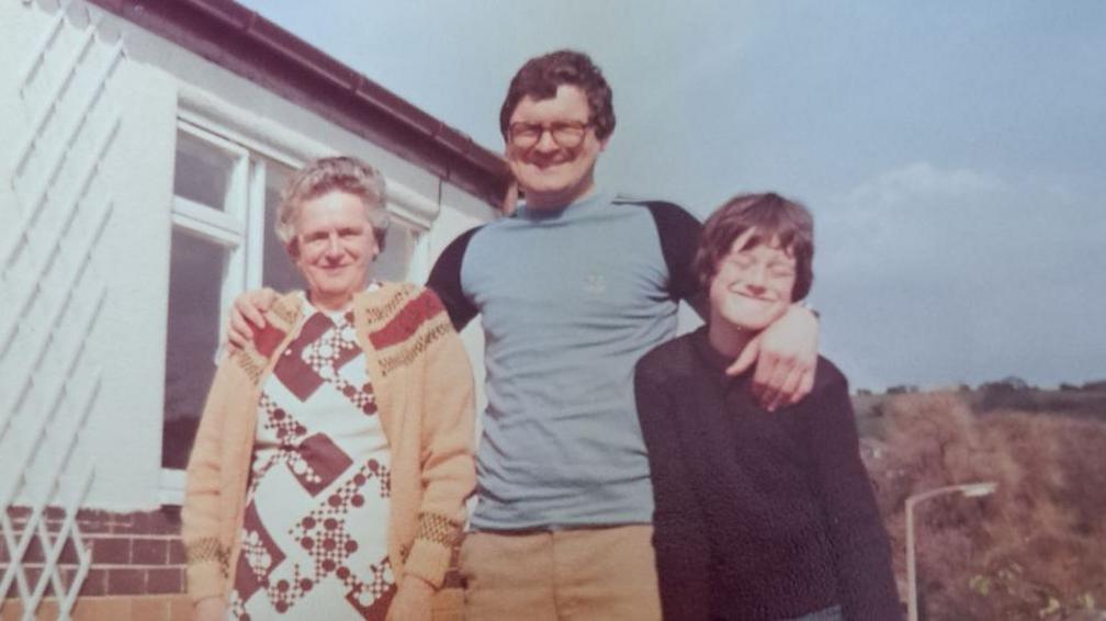 Kenneth is in the middle of the photograph with his arm around his mum on his right and his son Alan on his left. Alan has his eyes closed in the picture and is smiling. Alan is wearing a dark long sleeve top and has brown hair. Margaret is smiling. She has grey hair and is wearing a mustard cardigan. Kenneth is wearing glasses, a sky blue top with dark sleeves and mustard trousers.