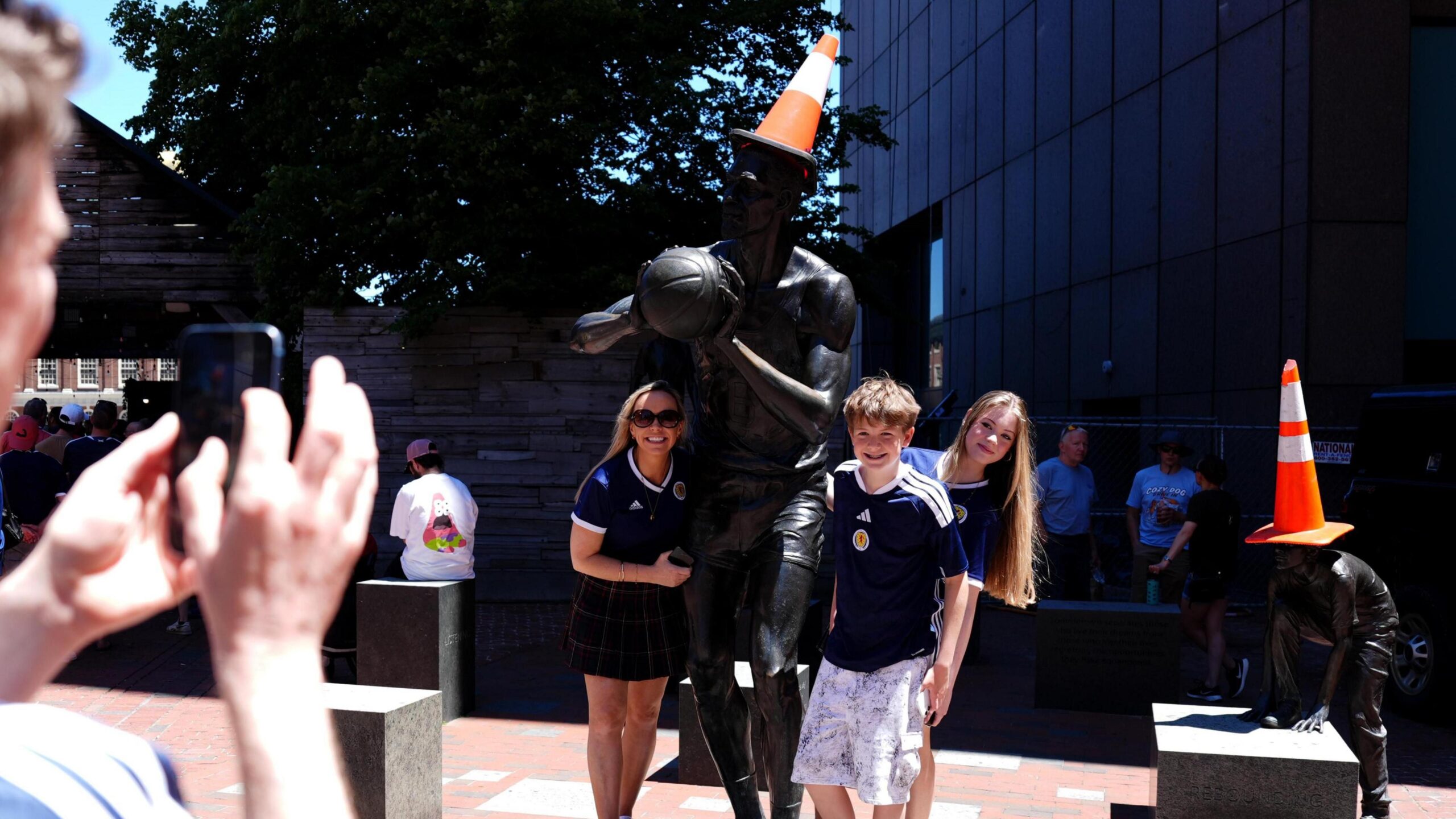 A man taking a photograph of two women and a boy wearing blue Scotland football shirts standing next to statues with orange traffic cones on their heads.